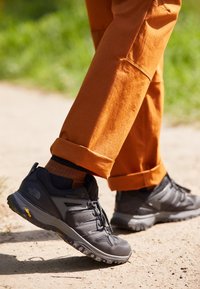 Un personne portant des chaussures de randonnée noires et un pantalon orange marchant sur un chemin de terre avec de l'herbe verte en arrière-plan.