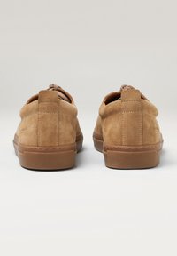 Zapatos de ante color arena con un perfil bajo, parte trasera redondeada y suela de goma texturizada. Presentan una costura y cordones marrones. Diseño limpio.