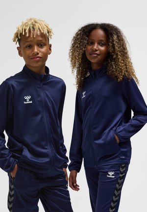 To børn iført marineblå Hummel sportjakker og bukser, stående side om side mod en ensfarvet baggrund.