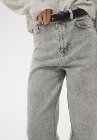 Ruka upravuje černý opasek na světle šedých džínách s vysokým pasem a jemnými třpytivými detaily, které jsou doplněny měkkým, huňatým světle šedým svetr.