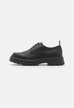 Chaussure basse à lacets en cuir noir avec semelle épaisse et robuste et boucle au talon, présentée sur fond blanc.