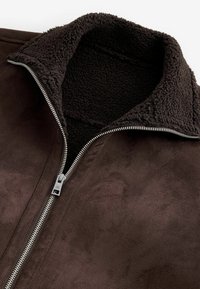Veste en daim marron avec une doublure en polaire douce marron foncé et une fermeture éclair argentée partiellement fermée au col avant.