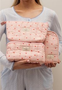 Lappede rosa kosmetikkvesker med røde hjerte-mønstre, med glidelåser og logo-tags. Tre størrelser stablet, holdt av en person.