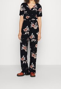 Kobieta w czarnej, kwiecistej bluzce z krótkim rękawem oraz pasujących szerokich spodniach, z brązowym obuwiem, stojąca na tle jasnym.