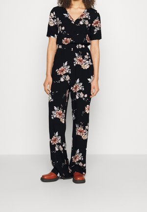 Mujer vestida con una blusa corta negra con flores y pantalones anchos a juego, con zapatos marrones, de pie frente a un fondo claro y liso.