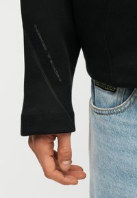 Χέρι χαλαρό στο πλάι, φοράει μαύρη μπλούζα με μακριά μανίκια και ραφές, και ανοιχτό μπλε τζιν με ορατή τσέπη και ετικέτα.