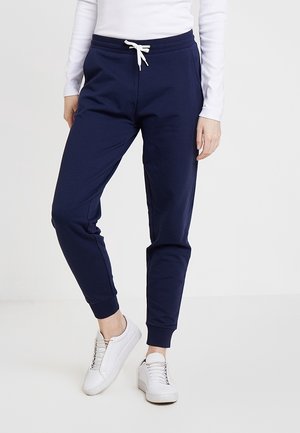 Personne portant un pantalon de jogging bleu marine avec des poignets élastiques et un cordon blanc, associé à des baskets blanches et un haut blanc à manches longues.