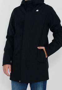 Veste noire à capuche avec une texture lisse, poches avant, design minimaliste et un petit logo sur la poitrine. Associée à un jean bleu.