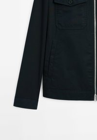 Manche de veste noire et bas du torse avec fermeture éclair frontale, poche à bouton et poche fendue latérale sur fond blanc.