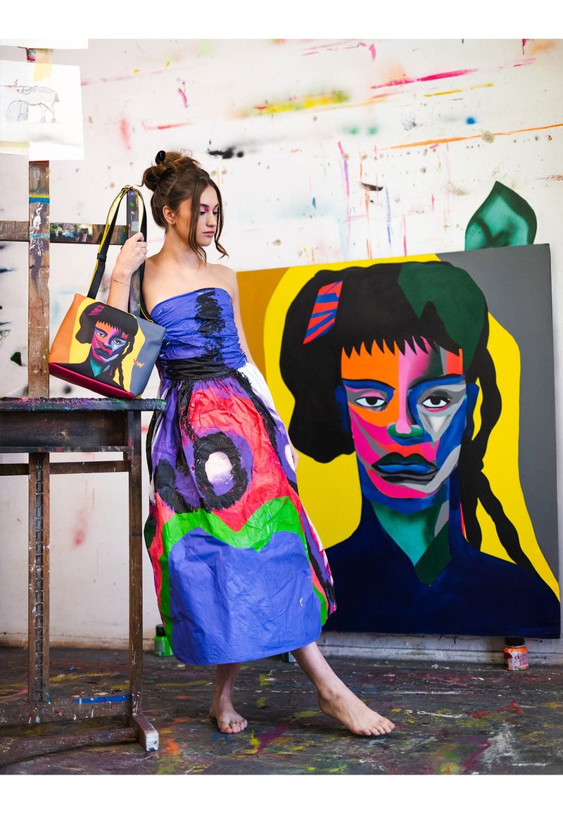 Buntes Kleid aus synthetischem Material, mit blauem Oberteil, schwarzen Akzenten und lebhaften Mustern. Das Model hält eine passende Handtasche.