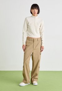 Crèmekleurige fleece half-zip pullover met lange mouwen, gecombineerd met losse beige cargobroeken met zijzakken. Een lichte achtergrond maakt de look compleet.