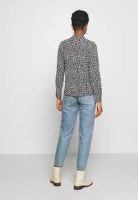 Långärmad blå blommig blus med nyckelhålsrygg, tillsammans med ljusblå jeans och krämfärgade ankelskor, i en neutral bakgrund.