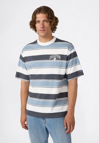 Man met krullend haar in een korteblauwe, witte en zwarte gestreepte Champion T-shirt en lichtblauwe jeans tegen een effen achtergrond.