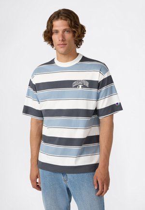 Man met krullend haar in een korteblauwe, witte en zwarte gestreepte Champion T-shirt en lichtblauwe jeans tegen een effen achtergrond.