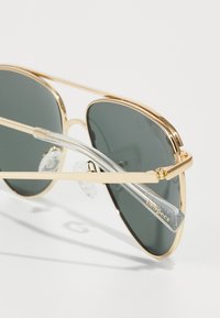 Guldmetall aviator-solglasögon med mörkgrå linser, klara nässtöd och minimalistisk ramdesign. Varumärket "Le Specs" är synligt på skalmen.