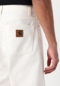 Hvide denimbukser har en baglomme med et brunt læderpatch med Carhartt-logoet. Stoffet har en blød tekstur med synlige syningdetaljer.