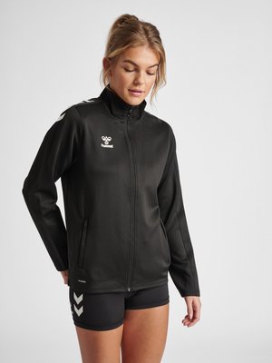 Femme en veste de sport noire zippée et short noir avec logos chevrons blancs, debout devant un fond clair uni, regardant légèrement vers le bas.