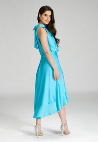 Bezramieniowa, jasnoniebieska sukienka o płynnej sylwetce, o satynowej fakturze, z przewiązanym akcentem z tyłu, zestawiona z srebrnymi sandałami na obcasie.