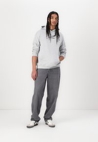 Siv hoodie z črnim logotipom, sive jeans hlače in bele teniske s črnimi črtami. Dolgi lasje, oblikovani v kite. Preprost, sproščen kroj.