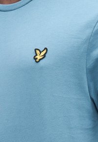 Light blue cotton t-shirt with a crew neck and short sleeves; features a small black and yellow embroidered eagle logo on the chest.