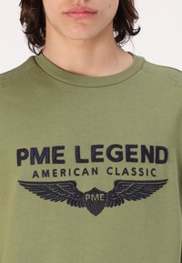 Maslinasto zeleni dukserica s rebrastim okovratnikom, na kojem se nalazi tamnoplavi vezeni natpis "PME LEGEND AMERICAN CLASSIC" i grafički naglasci s krilima.