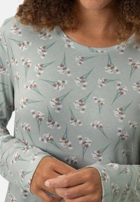 Jasnozielona bluzka z długim rękawem z kwiatowym wzorem w białe kwiaty z brązowym środkiem. Miękka tkanina z okrągłym dekoltem i dopasowanymi mankietami.