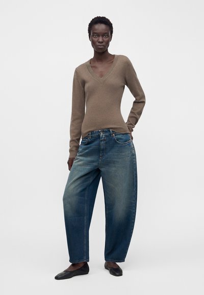 Femme debout portant un pull marron à col en V, un jean bleu délavé ample et des ballerines noires sur un fond clair uni.