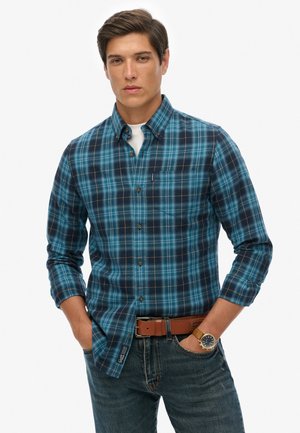 Jeune homme portant une chemise à carreaux bleue et noire boutonnée, un jean bleu, une ceinture marron et une montre-bracelet, debout les mains dans les poches.