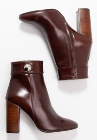 Botas de cuero marrón hasta el tobillo con punta afilada, que cuentan con un zipper en el lateral y un tacón de madera; acentuadas con un botón metálico circular.