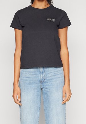 Kobieta ubrana w czarny krótki t-shirt Levi's i jasnoniebieskie wysokie jeansy, stojąca na jednolitym tle.