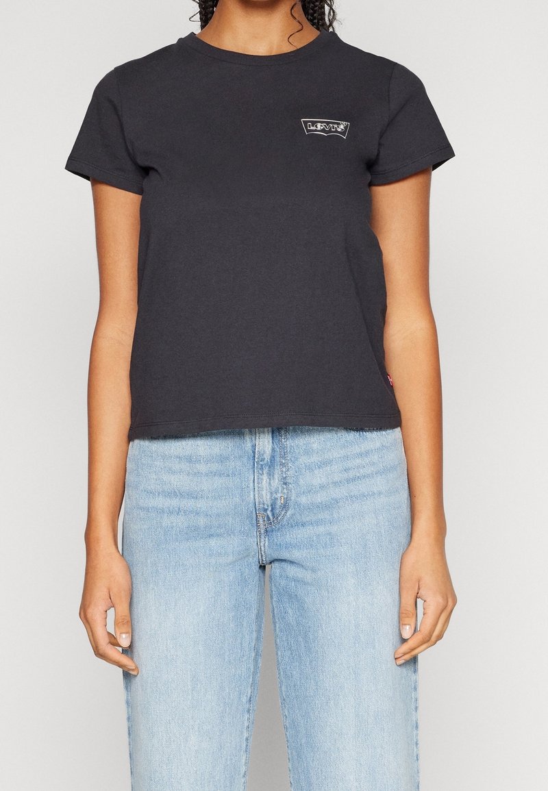 Femme portant un t-shirt court noir Levi's et un jean taille haute bleu clair, debout devant un fond uni.