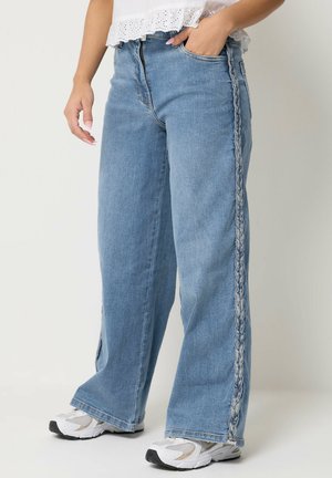 Personne portant un jean large bleu clair avec des coutures tressées extérieures, des baskets blanches et un crop top blanc en broderie anglaise.