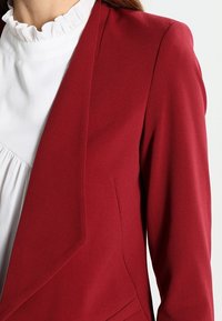 Blazer rosso con una vestibilità su misura, dotato di revers a punta e texture liscia, abbinato a una blusa bianca con volant.