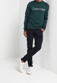 Sudadera verde de cuello redondo con el texto blanco "Calvin Klein", combinada con pantalones oscuros y zapatillas blancas, de pie frente a un fondo neutro.