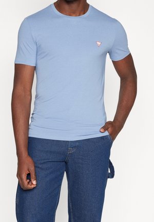 Mężczyzna ubrany w jasnoniebieską koszulkę z krótkim rękawem z małym czerwonym trójkątnym logo na piersi, w połączeniu z ciemnoniebieskimi dżinsami.