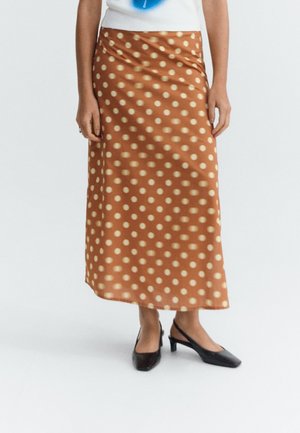 Femme portant une jupe midi marron à pois beiges et des chaussures à bride arrière noires à talons bas, debout devant un fond uni.