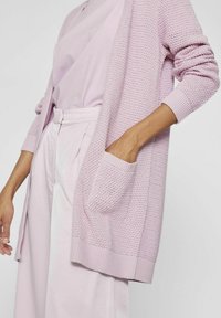 Cardigan lavorato a maglia rosa chiaro con un design testurizzato, polsini a coste e una tasca frontale. Indossato sopra un top e pantaloni abbinati.