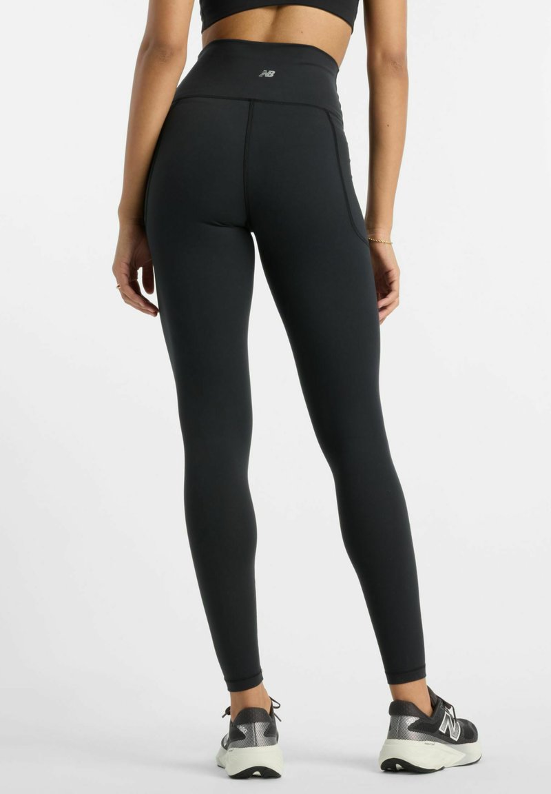 Leggings noirs taille haute en tissu extensible. Caractérisés par une texture lisse avec des poches latérales, accompagnés de baskets grises et noires.