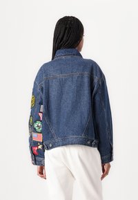 Jeansjacka med avslappnad passform, blåtvätt, klassisk krage och olika färgglada lappar på vänstra ärmen.