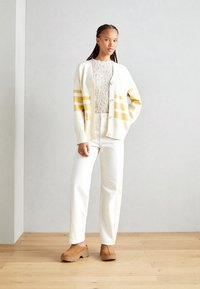 Cardigan bianco con strisce gialle, indossato su una maglia bianca con texture, abbinato a pantaloni bianchi e scarpe con zeppa marroni su un pavimento di legno.