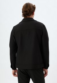 Sort ribbet jakke med krave, tekstureret stof og en afslappet pasform. Bagperspektivet viser skuldersømme og manchetter med minimal detaljering.