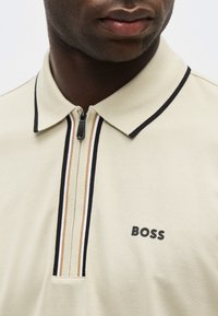 Άνδρας που φοράει μπεζ πόλο με φερμουάρ, με ίσιες ρίγες σε μαύρο, μπεζ και άσπρο στο φερμουάρ και μαύρο λογότυπο "BOSS" στο στήθος.