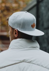 Cappellino da baseball grigio con una toppa in pelle marrone, indossato al contrario, abbinato a un giubbotto bianco imbottito. Lo sfondo presenta un fogliame autunnale sfocato.