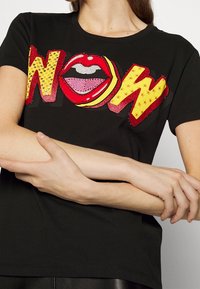 Černé tričko s nápisem "WOW" a grafikou červených rtů, zdobené flitry v různých barvách. Krátké rukávy a kulatý výstřih.