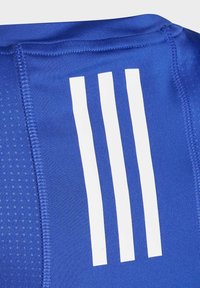 Camiseta deportiva azul con tejido texturizado, franjas blancas contrastantes y costuras visibles. El diseño enfatiza la funcionalidad y el espíritu deportivo.
