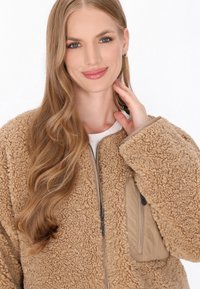 Beige Fleecejacke mit strukturiertem, flauschigem Außenmaterial, Reißverschluss vorne und Seitentasche. Weicher Stoff mit lockerer Passform und weiten Ärmeln.