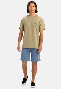 Camiseta de algodón verde oliva claro con un pequeño logo negro, combinada con pantalones cortos azules claros que tienen un parche de logo circular, complementando zapatillas negras sin cordones.