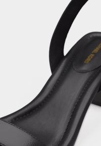 Κοντινό πλάνο μαύρου δερμάτινου σανδαλιού με επενδεδυμένη σόλα και λουράκι στον αστράγαλο, που αναδεικνύει το ράψιμο και την ομαλή υφή.