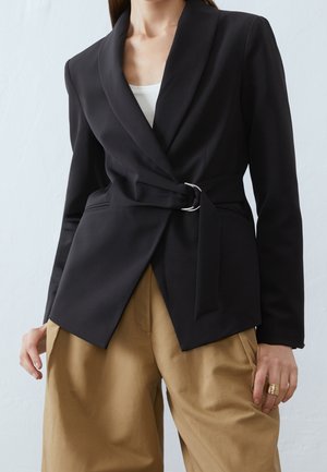 Femme portant un blazer noir cintré à porter façon portefeuille avec une boucle argentée, un haut blanc et un pantalon beige ample, debout devant un fond clair.