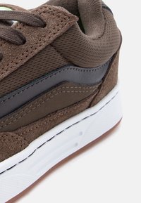 Καφέ τακούνι αθλητικού παπουτσιού με λεπτομέρειες από σουέτ και δίχτυ, μαύρη δερμάτινη λωρίδα και λευκή υφασμάτινη σόλα με καφέ λαστιχένιο κάτω μέρος.